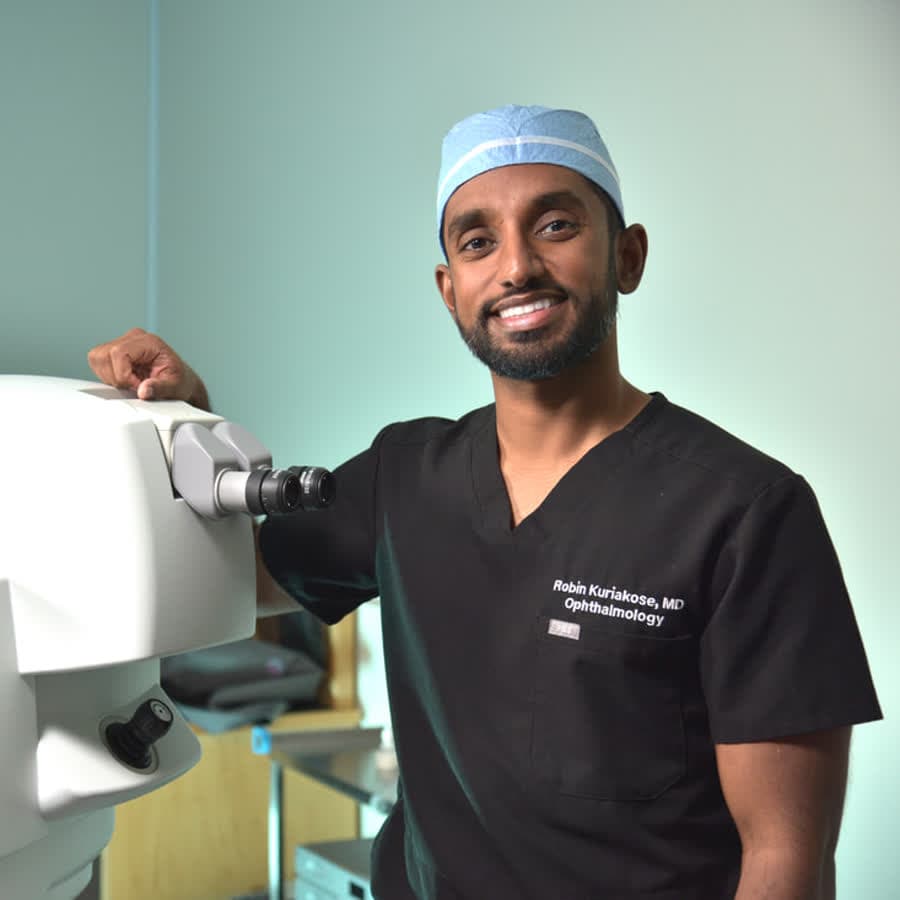 Dr. Robin Kuriakose, MD, in scrubs and a surgical cap beside a laser at Optima Eye in Hayward, California.
